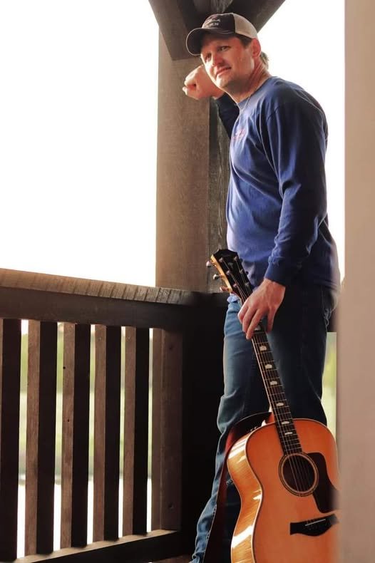 Brett Sanders standing with an acoustic guitar on a wooden balcony, promoting North Florida country and acoustic live music performances.