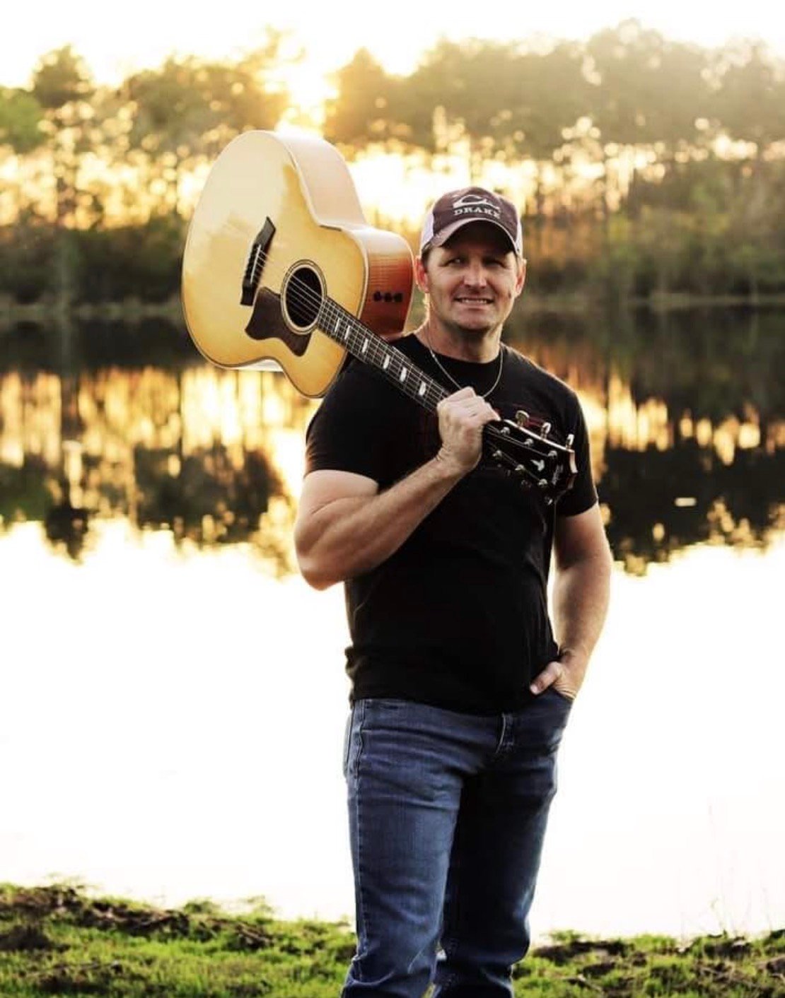 Brent Saunders, a North Florida solo acoustic performer, holding a guitar beside a lake at sunset